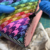 Close-up of blue-gloved hands applying black adhesive with a small brush along the stitched edge of a colorful quilted sneaker upper.